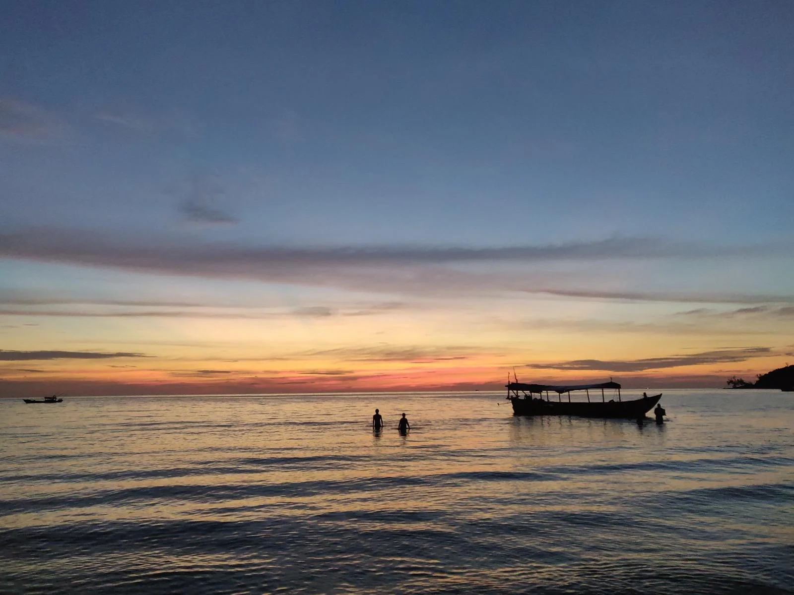 Sunset over the sea off Koh Rong Sanloem, a wooden boat at anchor and two people standing in the shallows
