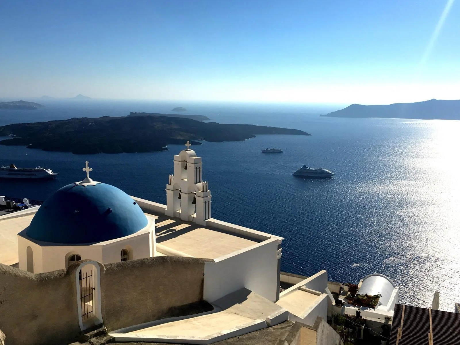 A blue-domed church and its bell tower above the caldera, cruise ships far below